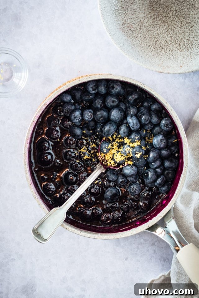 Overhead shot of lemon zest being stirred into the warm blueberry sauce in a pan, along with additional fresh berries.