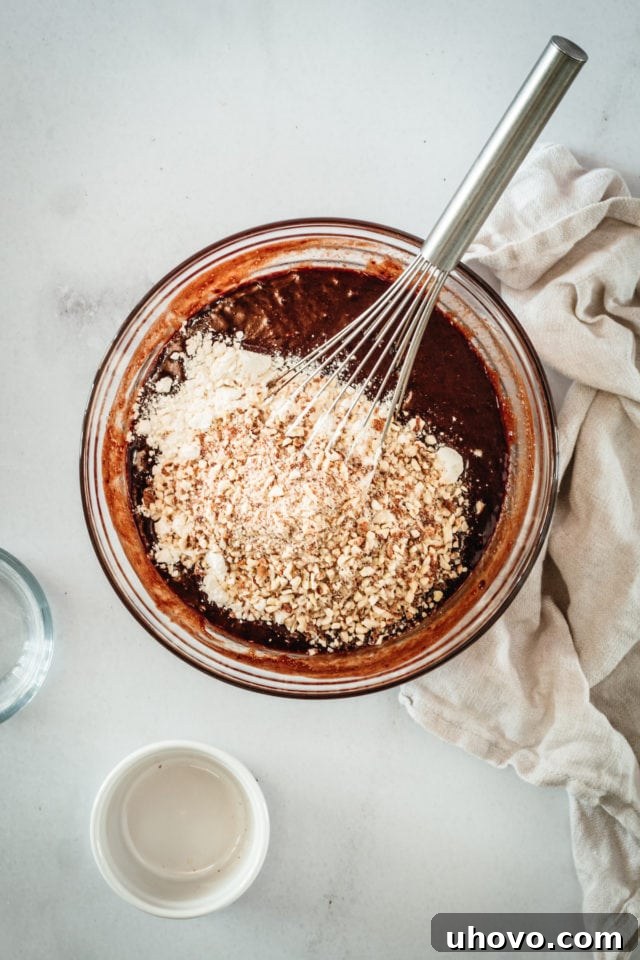 Overhead view of dry ingredients, including flour and nuts, being gently folded into the luscious brownie batter