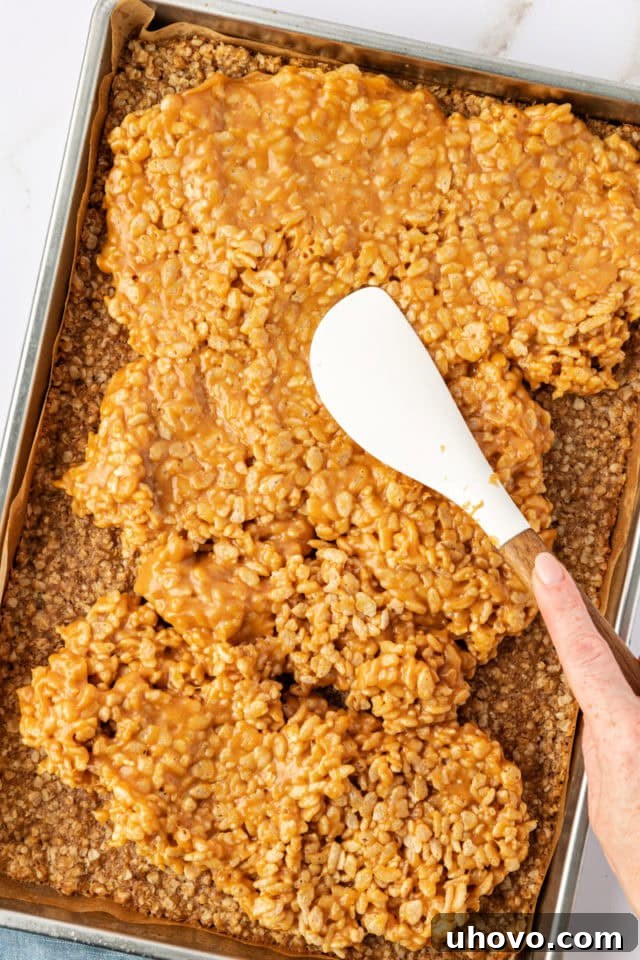 The peanut butter cereal filling being carefully spread over the slightly cooled oat crust in the jelly roll pan, creating an even layer.