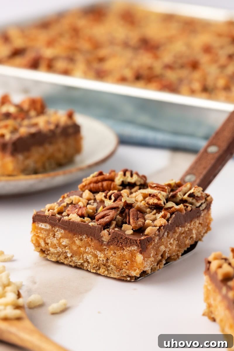 A single chocolate peanut butter cereal bar perfectly balanced on a cookie spatula, with a blurred background of more bars in the pan, ready to be served.