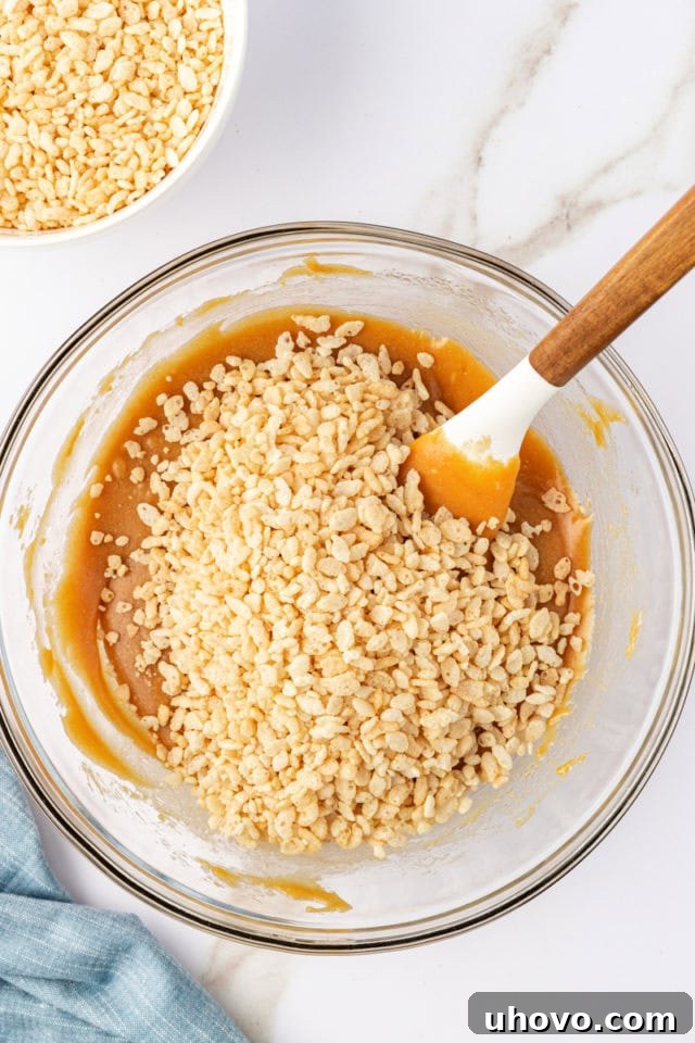 Crisp rice cereal being added to the melted peanut butter mixture in a glass bowl, creating the second layer of the bars.