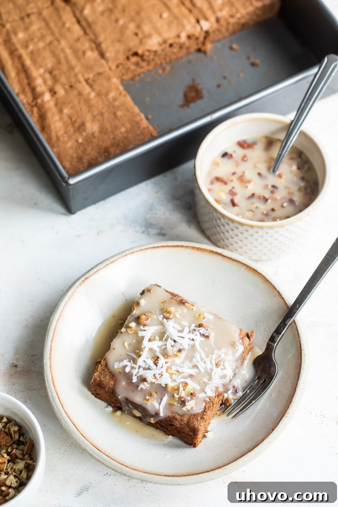 An inviting closeup of a square of German chocolate cake, topped with a rich, shimmering coconut-pecan sauce, resting on a beige plate with a fork nearby.