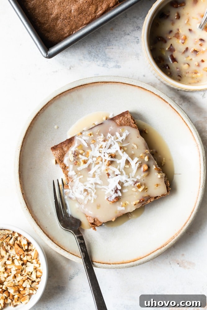 An overhead shot of a freshly baked German chocolate cake square, generously topped with its signature warm coconut-pecan sauce, resting on a beige plate. The cake's surface glistens with the sweet, nutty topping.