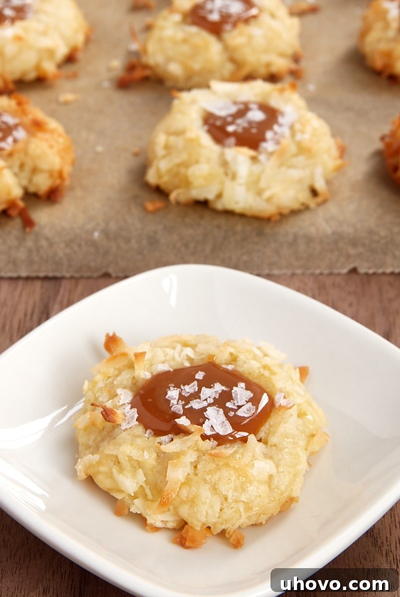A single Coconut-Salted Caramel Thumbprint Cookie on a white plate, with more cookies blurring in the background.