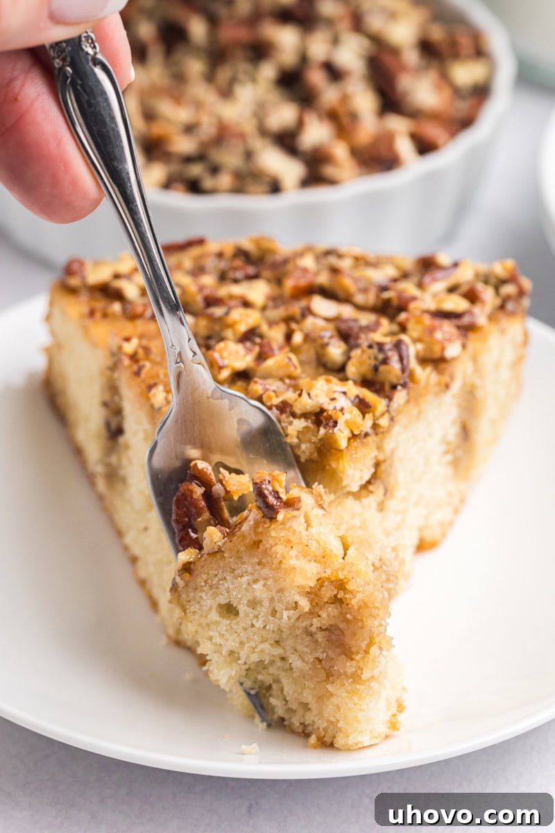 A fork gently cuts into a perfectly baked slice of Pecan Pie Coffee Cake on a white plate, showcasing its tender layers and rich topping.