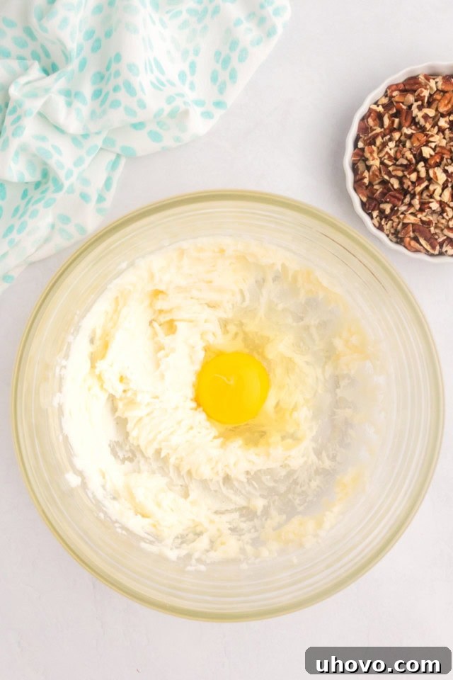 An overhead shot showing a cracked egg being added to a glass mixing bowl, already containing creamy, whipped butter, cream cheese, and sugar mixture, poised for the next step of the coffee cake batter preparation.