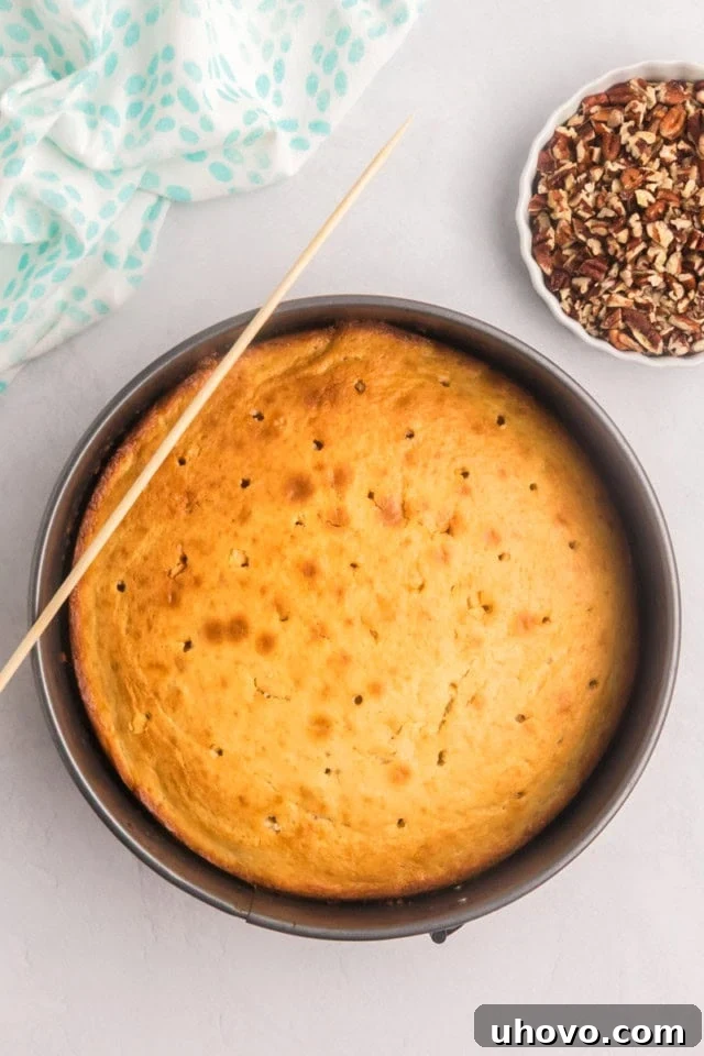 An overhead shot of a freshly baked pecan pie coffee cake, still in its pan, with a skewer used to poke numerous holes across its surface, preparing it for the topping.