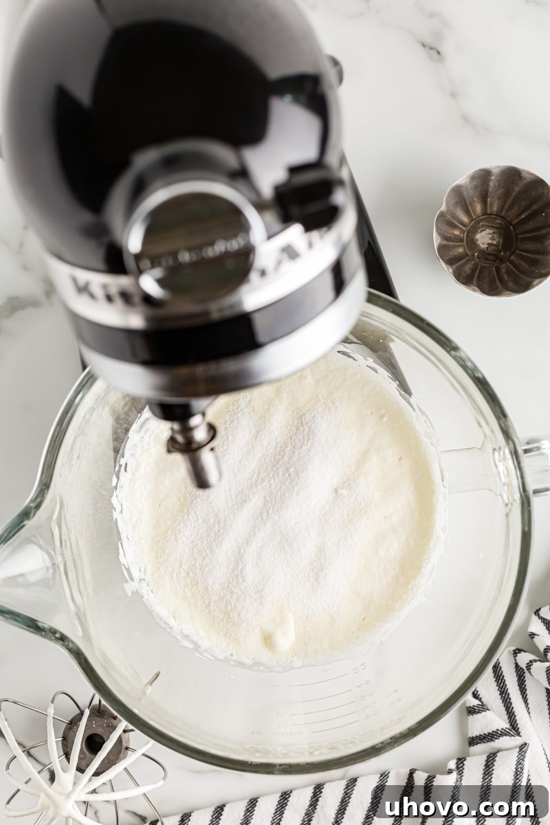 Overhead view of heavy cream with granulated sugar sprinkled over it in a stand mixer bowl, continuing to be whipped.
