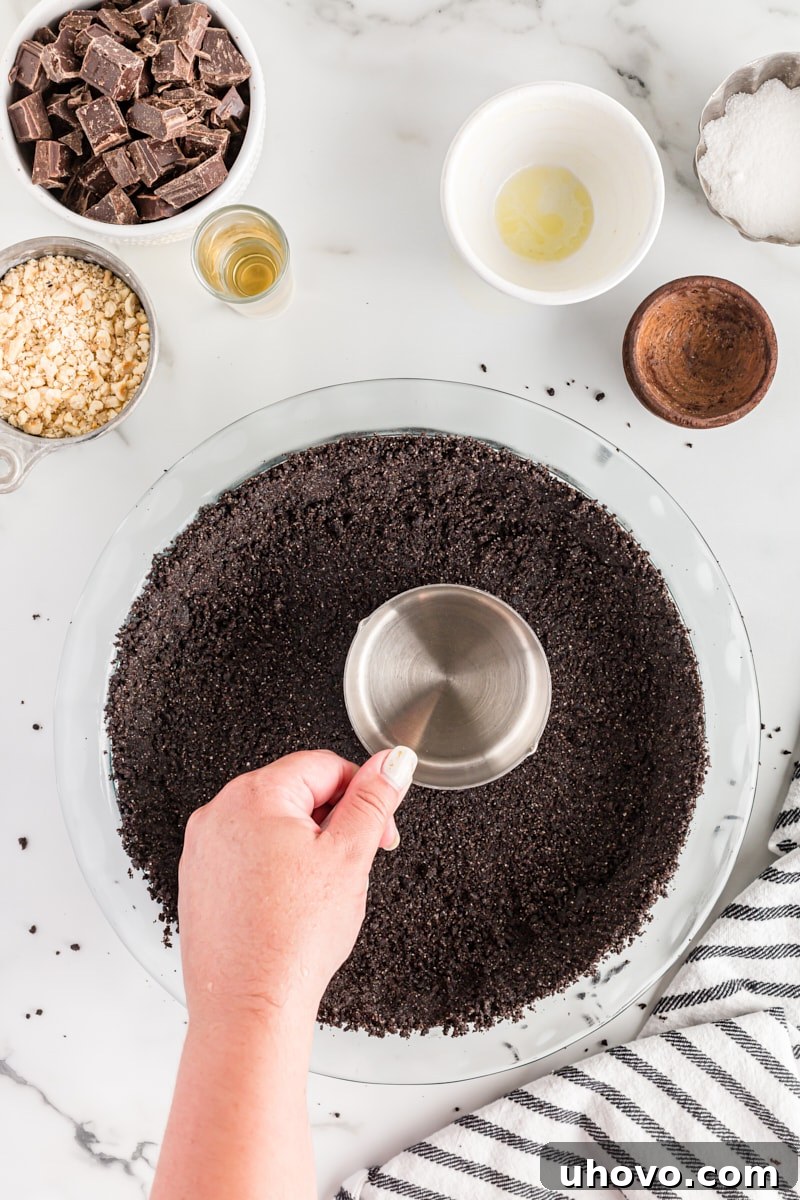 An overhead shot of a measuring cup being used to firmly press the chocolate cookie crust mixture into a pie plate.