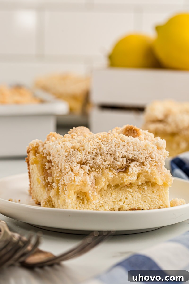 Lemon yogurt crumb cake piece on plate with rest of cake and lemons in background