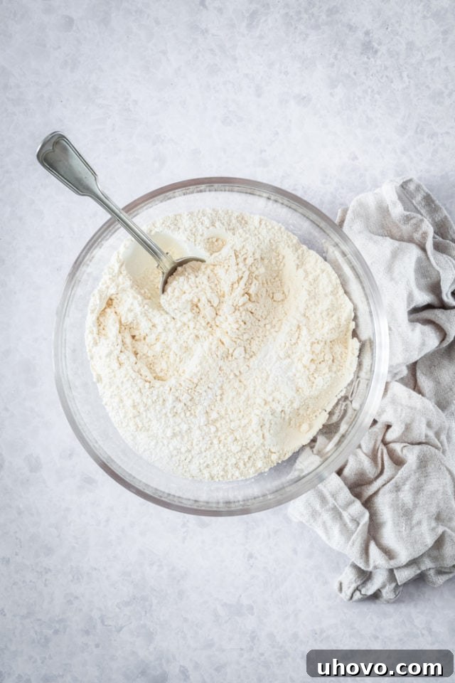 Overhead view of dry ingredients - flour, baking powder, baking soda, and salt - in a clear glass mixing bowl, ready to be combined.