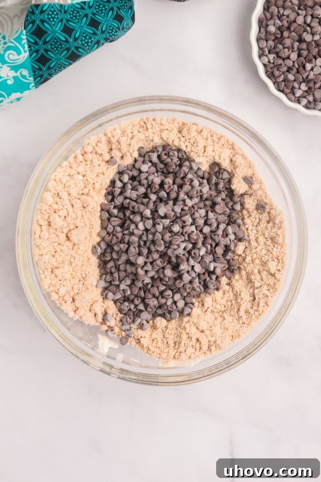 An overhead shot showing a generous handful of mini chocolate chips being added to the crumb topping mixture in a glass bowl, ready to be incorporated.
