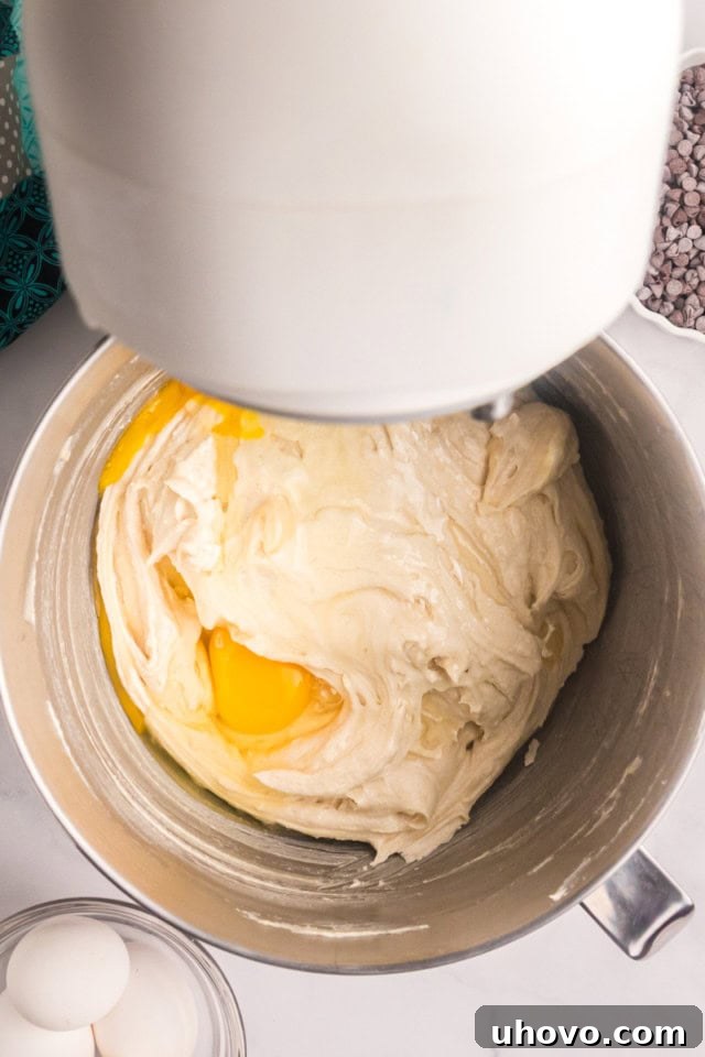 A vibrant overhead shot of eggs being added, two at a time, to the rich, creamed butter and sugar mixture within the bowl of a stand mixer.