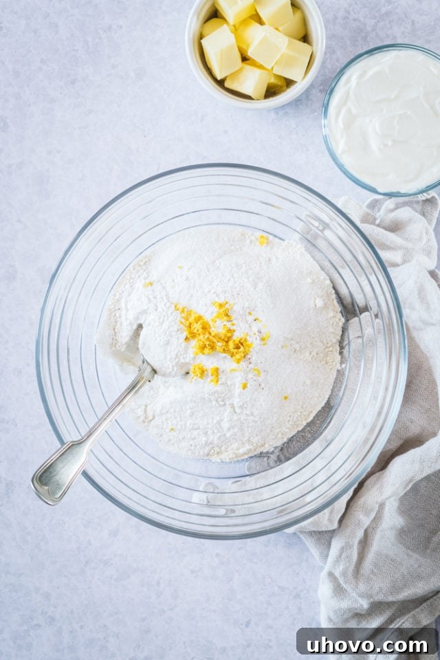 Overhead view of a glass mixing bowl containing flour, sugar, baking powder, lemon zest, and salt, perfectly combined.