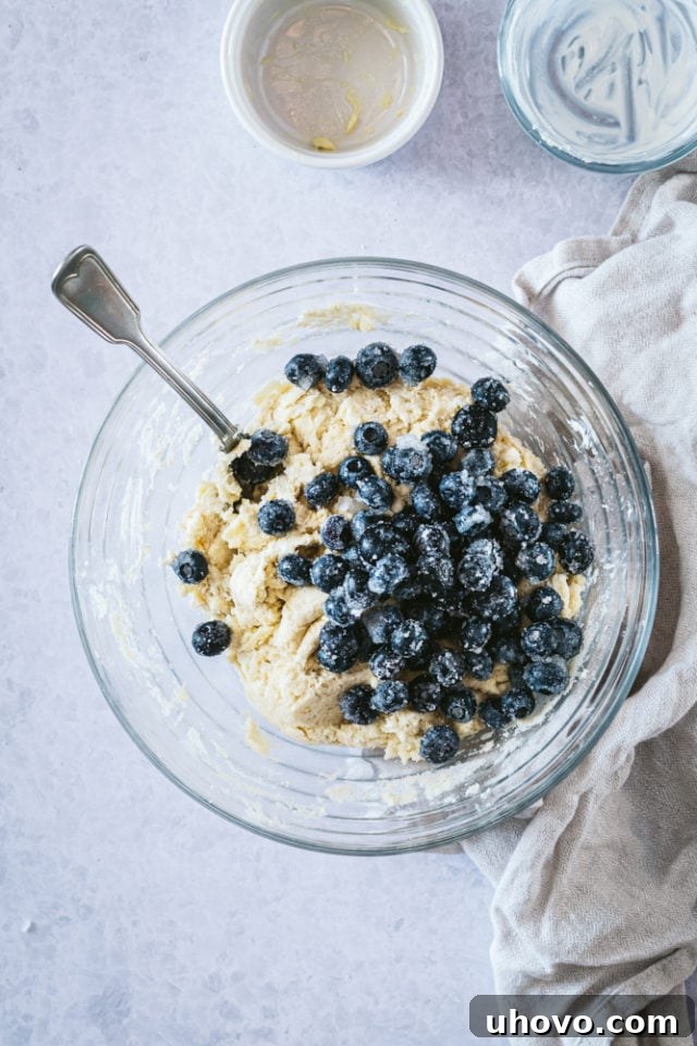 Overhead view of the blueberry mixture being gently folded into the cookie dough in a glass mixing bowl.
