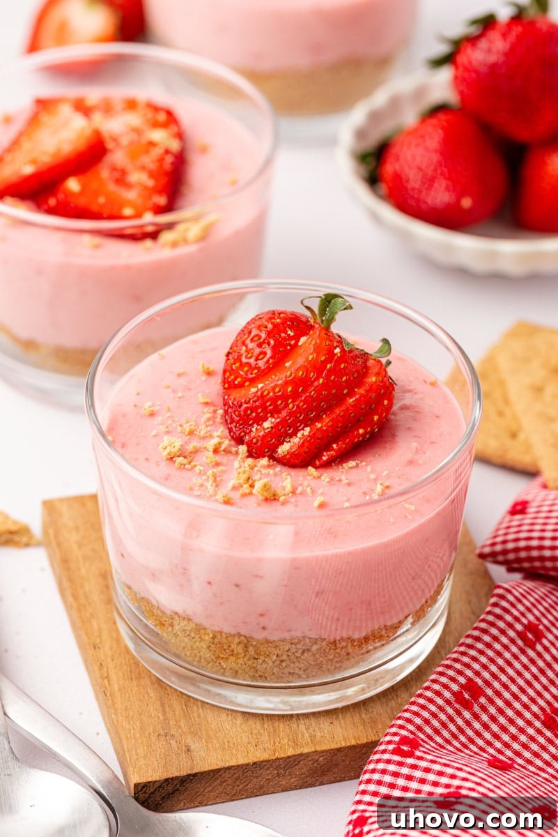 A beautifully presented individual strawberry no-bake cheesecake in a clear glass, with a soft focus background showing more cheesecakes and a bowl of fresh strawberries.