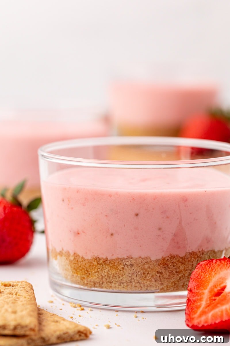 A close-up view of a single strawberry no-bake cheesecake in a clear glass, showing its distinct layers of graham cracker crust and creamy pink filling, with more cheesecakes subtly blurred in the background.
