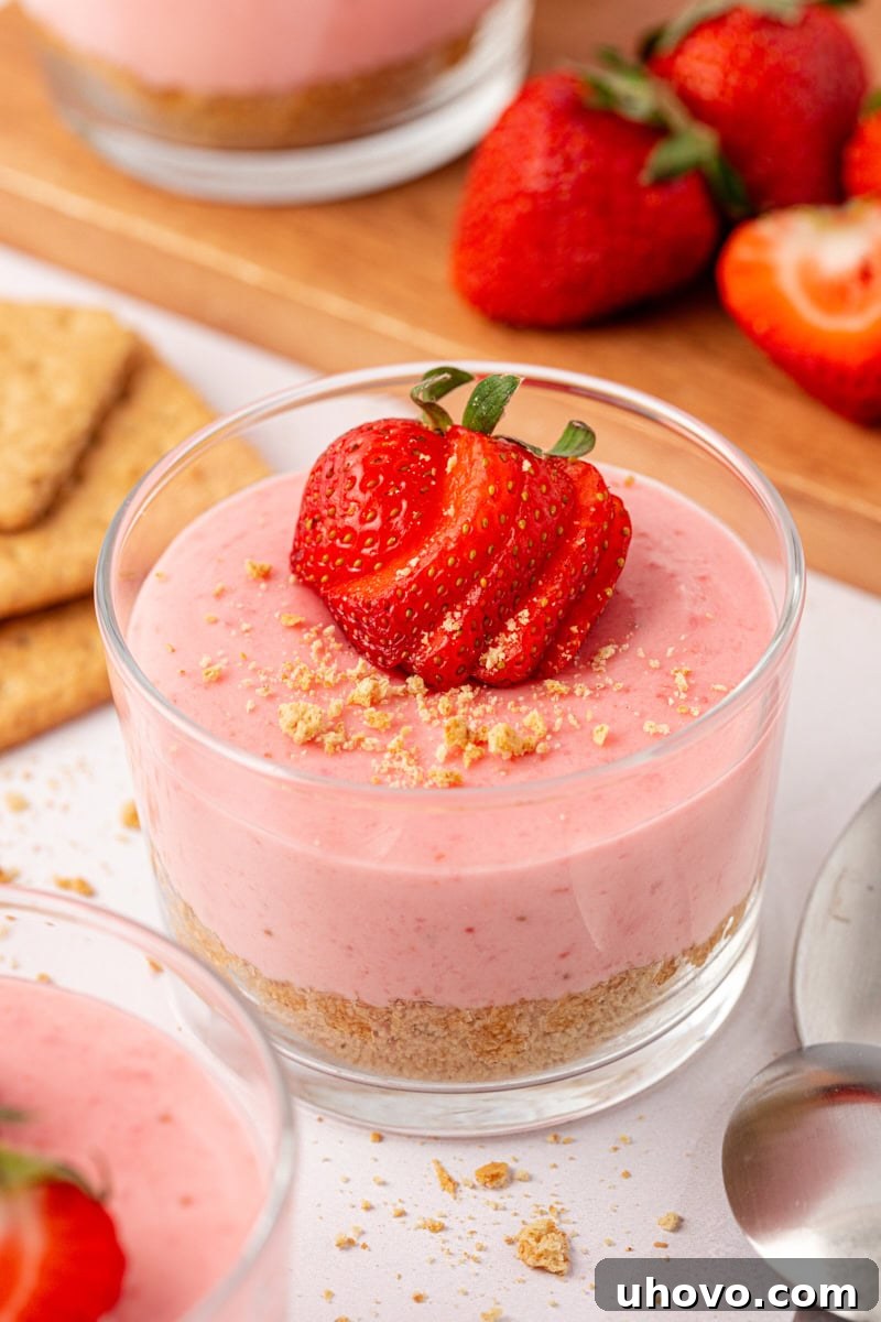 A vibrant strawberry no-bake cheesecake, generously topped with fresh strawberry slices and a light scattering of graham cracker crumbs, positioned against a soft, blurred background of more desserts.
