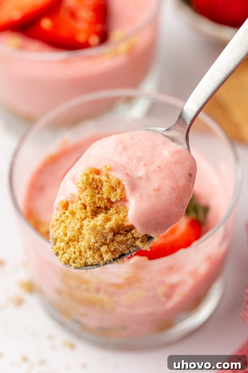 A close-up shot of a spoon taking a generous bite from a strawberry no-bake cheesecake, highlighting the creamy texture and crumbly base, with the remaining cheesecake in a glass blurred in the background.