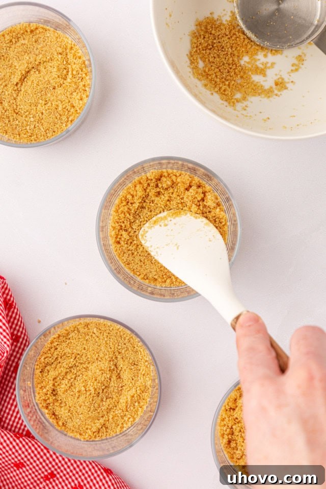 Close-up overhead view of graham cracker crumbs being pressed firmly into the bottom of a small, clear glass, forming the cheesecake crust.