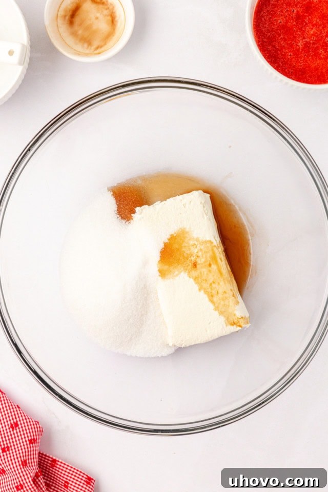 A clear glass mixing bowl containing softened cream cheese, granulated sugar, and a splash of vanilla extract, ready to be beaten until smooth.