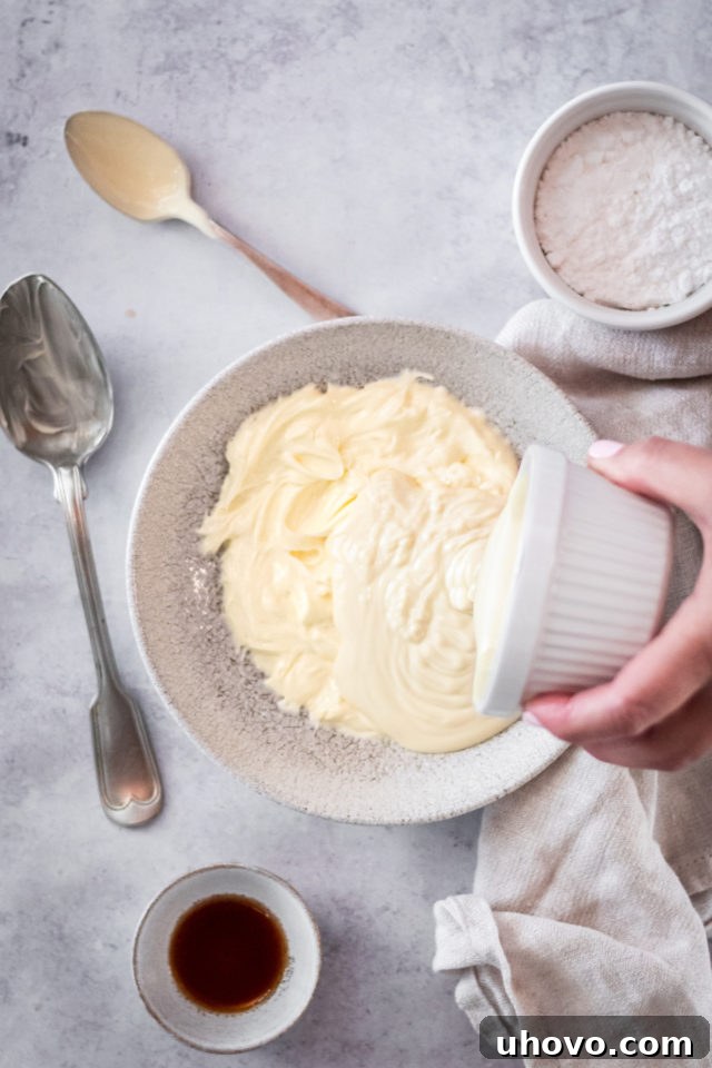Overhead shot of a hand pouring melted white chocolate into a bowl of creamed butter, ready to be mixed for frosting.