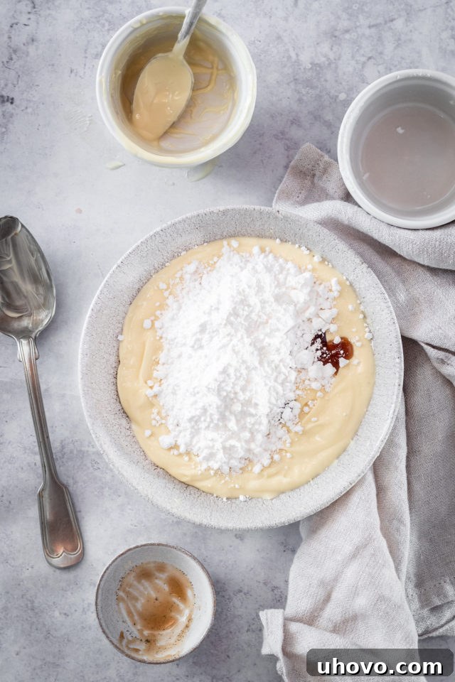 Overhead process shot of confectioners' sugar and vanilla extract being added to the white chocolate and butter mixture for frosting.