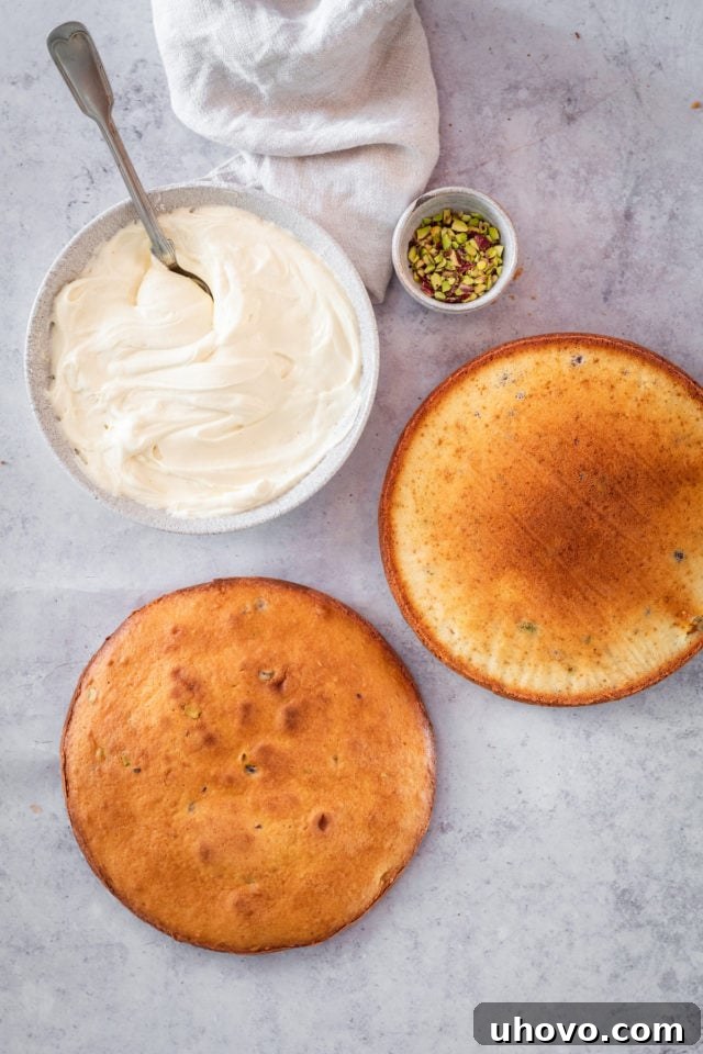 Overhead shot of two cooled, unfrosted cake layers, a bowl of white chocolate frosting, and a small bowl of chopped pistachios, ready for assembly.