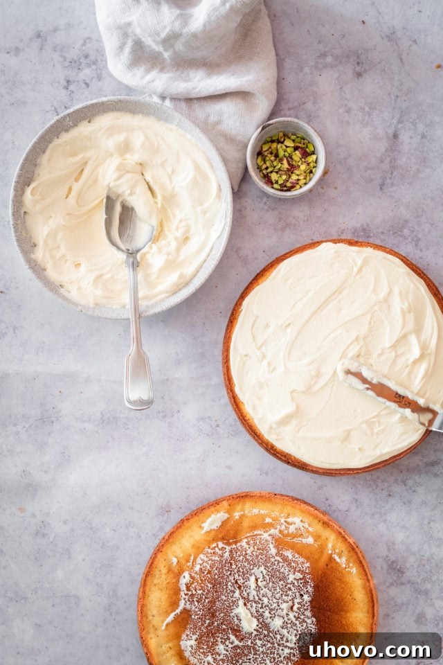 Overhead process shot of a cake layer being frosted with white chocolate buttercream using an offset spatula on a cake stand.