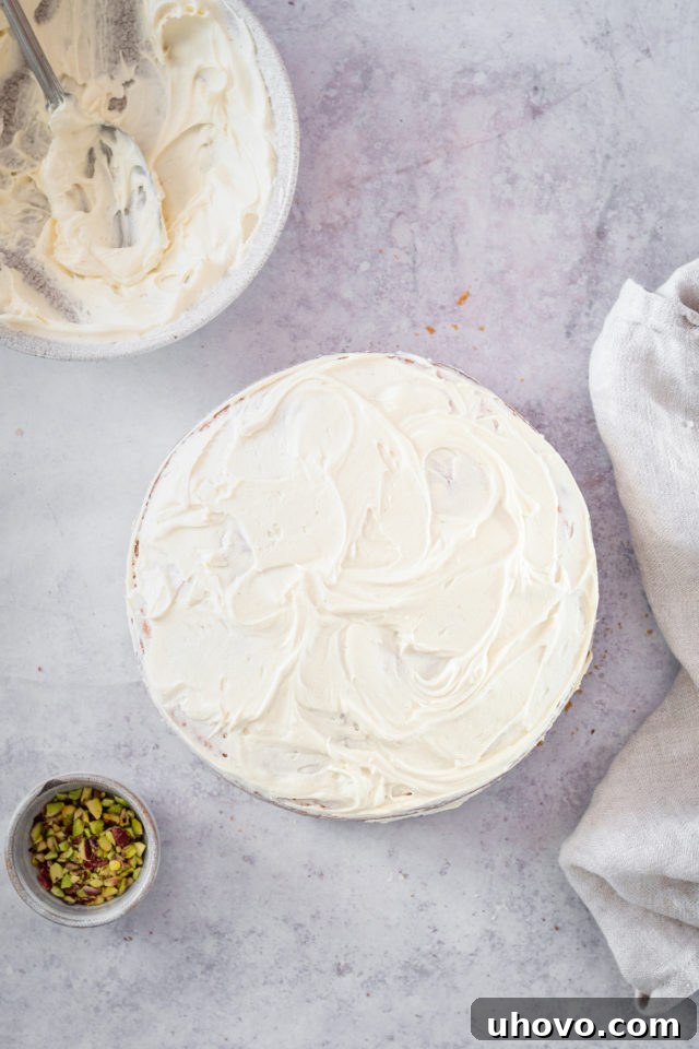 Overhead view of a two-layer frosted cake with bowls of remaining white chocolate frosting and chopped pistachios in the background.