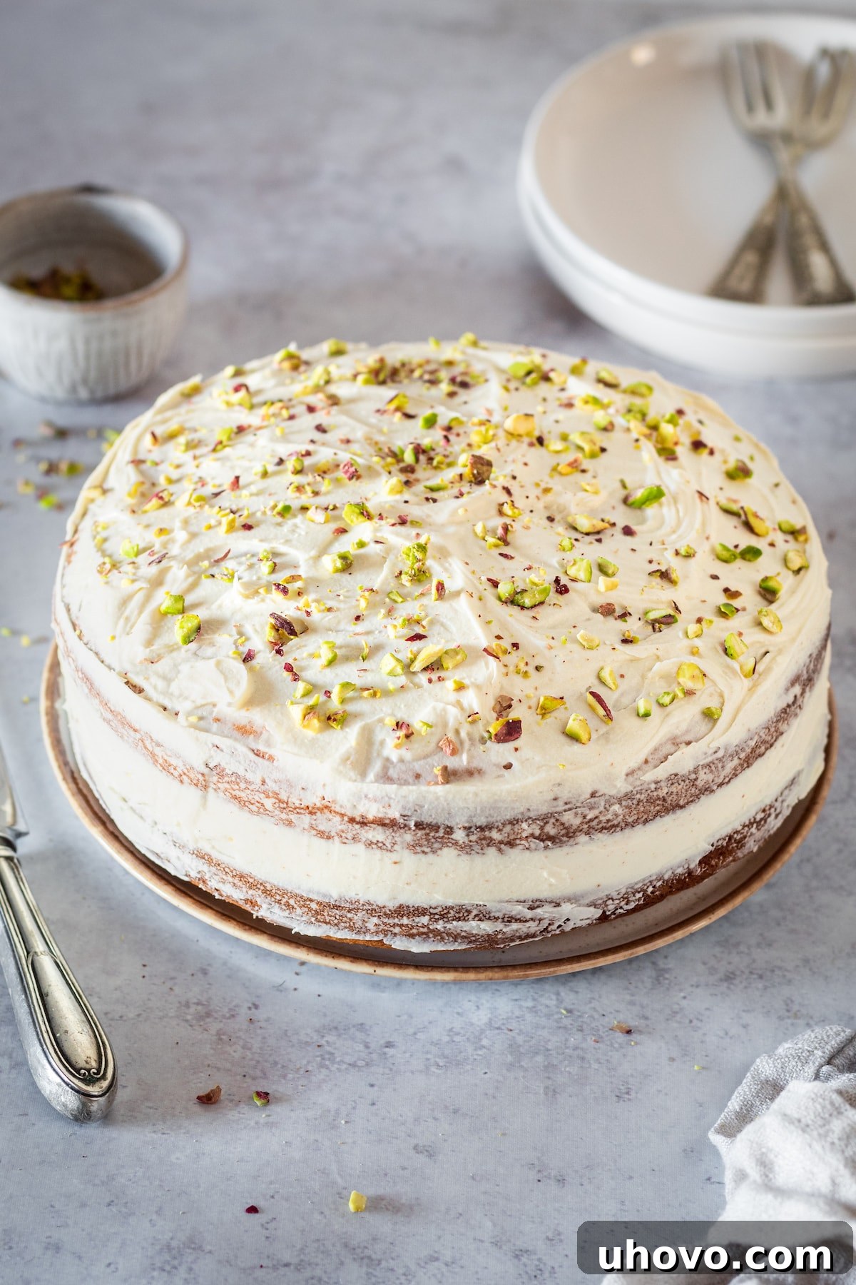 Whole Pistachio Cake beautifully frosted with white chocolate buttercream and garnished with pistachios, with serving plates and forks in the background.