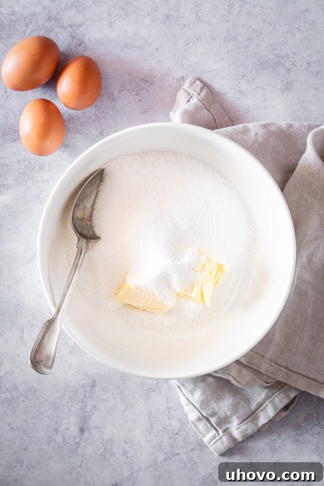 Overhead shot of softened butter and granulated sugar being creamed together in a large white bowl with an electric mixer, with whole eggs in the background.