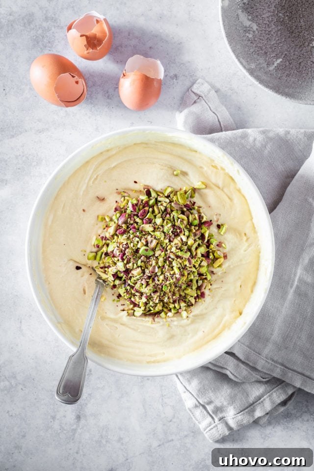 Overhead shot of chopped pistachios being folded into the cake batter in a large bowl, with a rubber spatula.