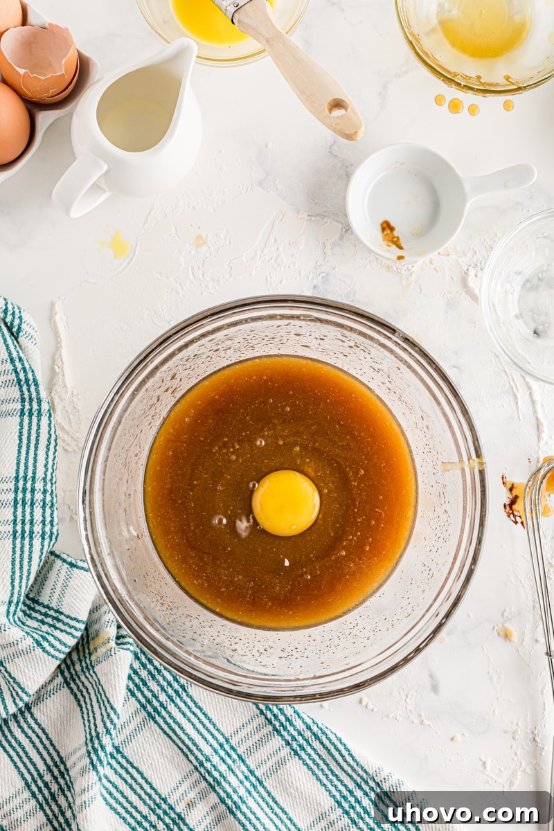 A large egg being cracked and added to a bowl containing a mixture of melted butter and brown sugar.