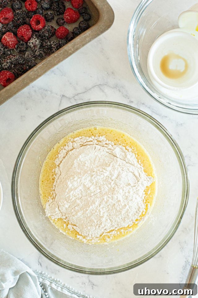 An overhead view of the dry flour mixture being gently incorporated into the wet egg and sugar mixture in a glass mixing bowl, signaling the start of batter formation.