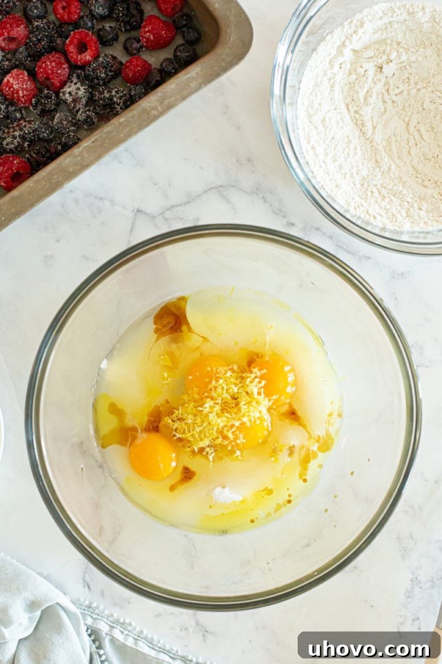 A pristine overhead shot of a bowl filled with the wet ingredients—granulated sugar, eggs, olive oil, fragrant lemon zest, and vanilla extract—all awaiting their turn to be mixed.