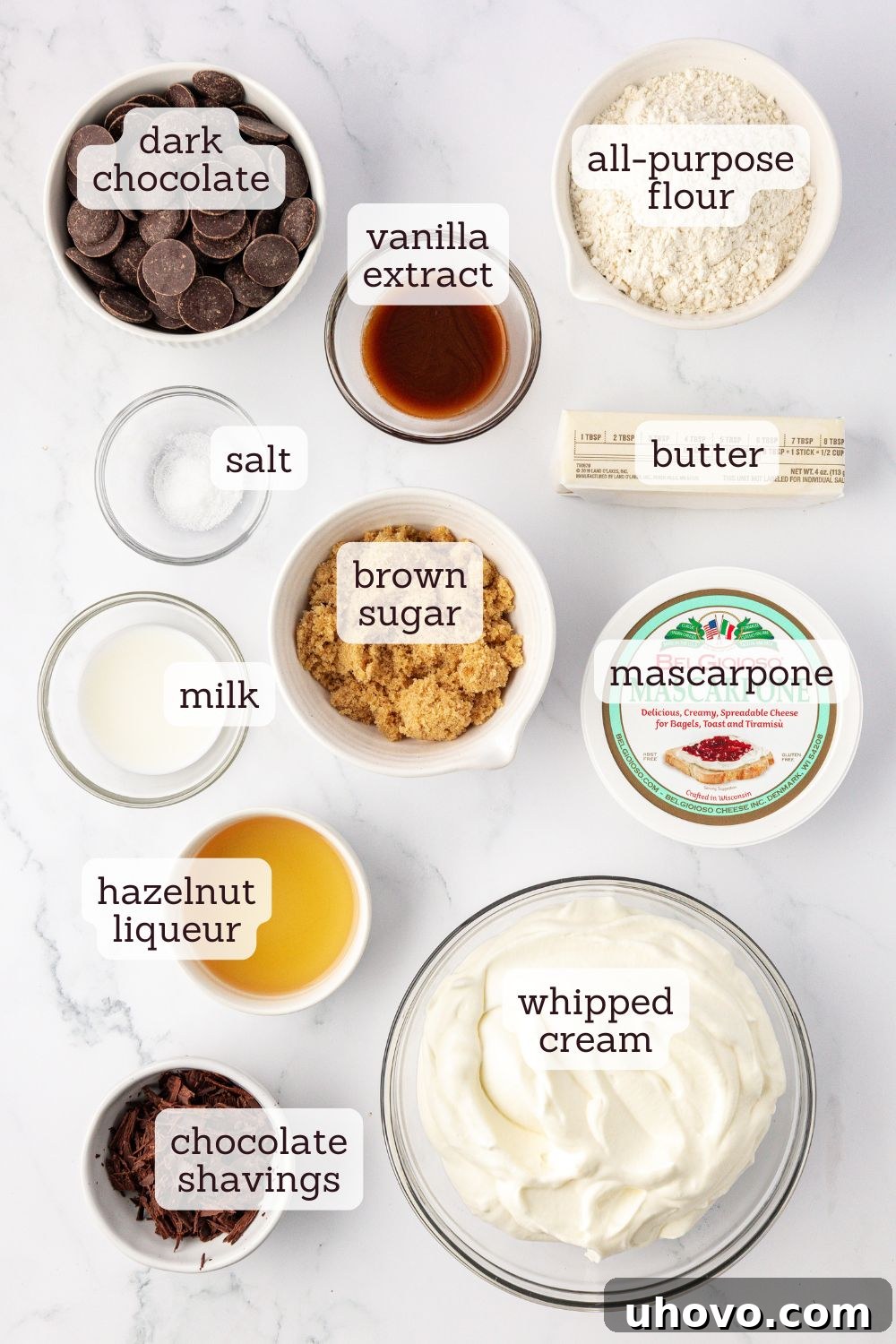 An overhead view of all the fresh ingredients meticulously laid out for a chocolate mascarpone tart, showcasing dark chocolate, mascarpone, butter, brown sugar, flour, and a bottle of hazelnut liqueur.