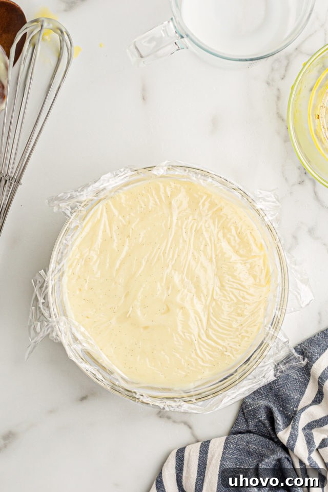 A glass bowl filled with warm vanilla pudding, meticulously covered with plastic wrap pressed directly onto its surface, ready for chilling.