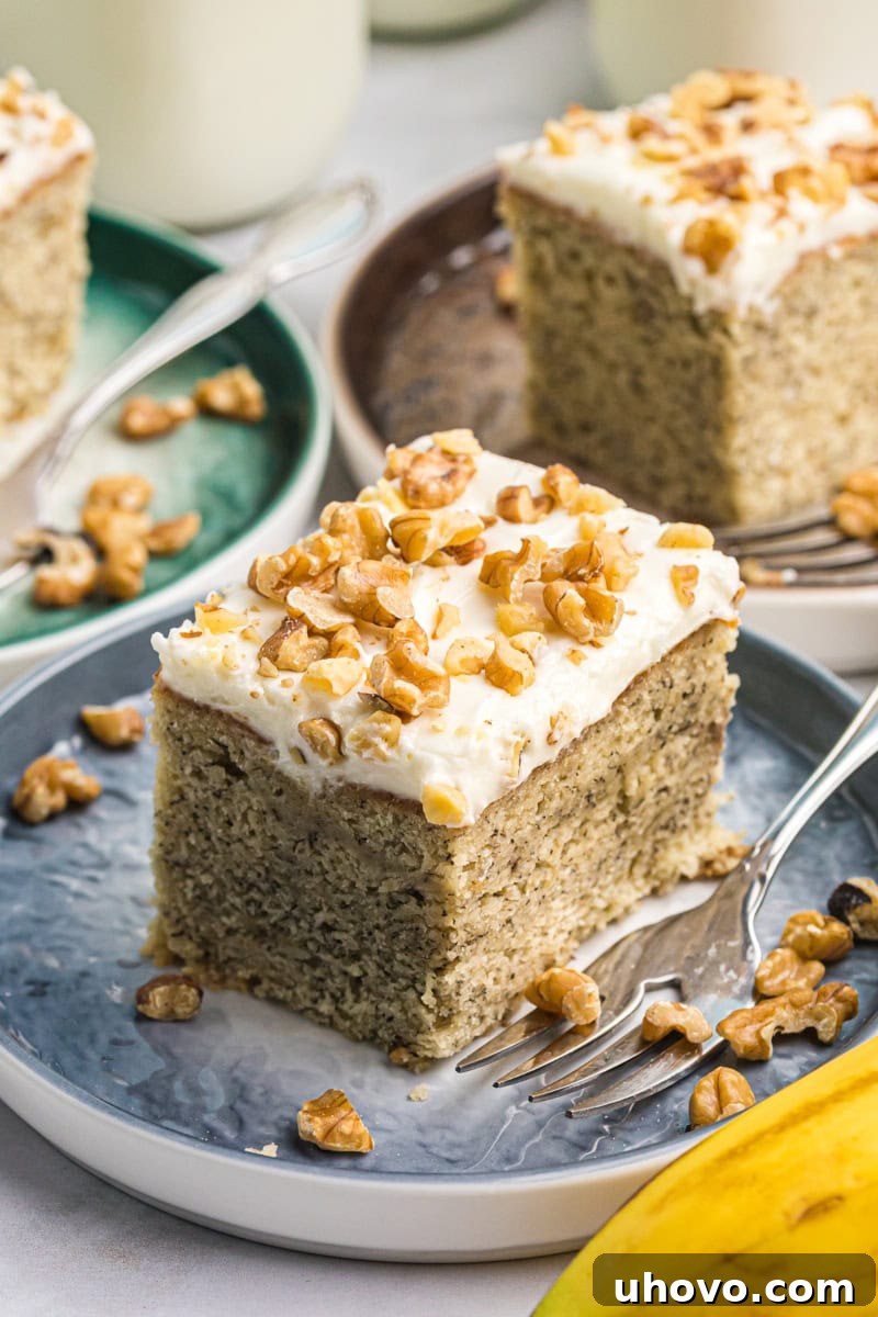 A perfectly sliced banana cake topped with rich cream cheese frosting, served on a blue plate. More frosted banana cake is visible in the background, hinting at its generous size.