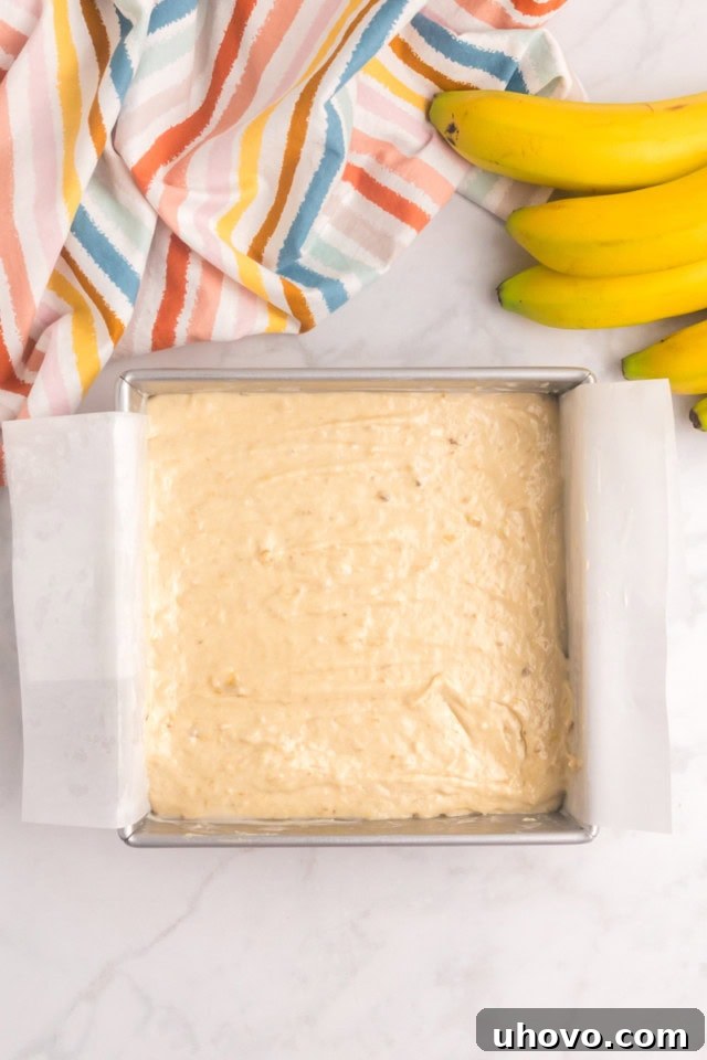 An overhead view of the banana cake batter smoothly spread into a parchment-lined 8-inch square baking pan, ready for the oven.