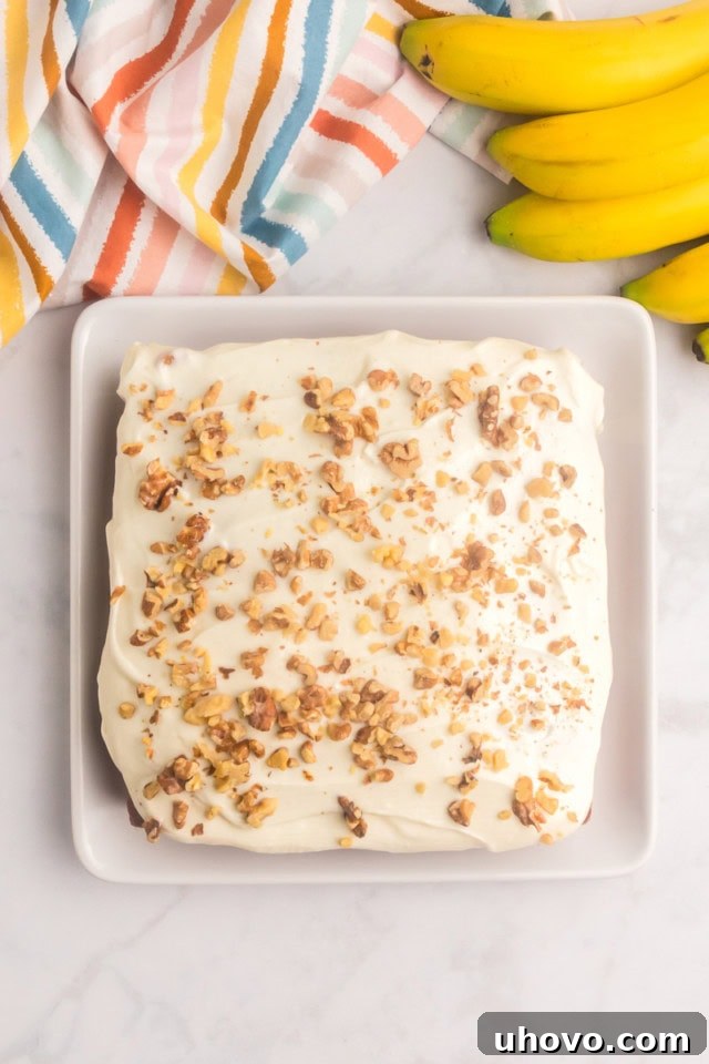 An overhead view of a beautifully frosted banana cake topped with toasted chopped nuts, with a slice served on a square white plate beside it.