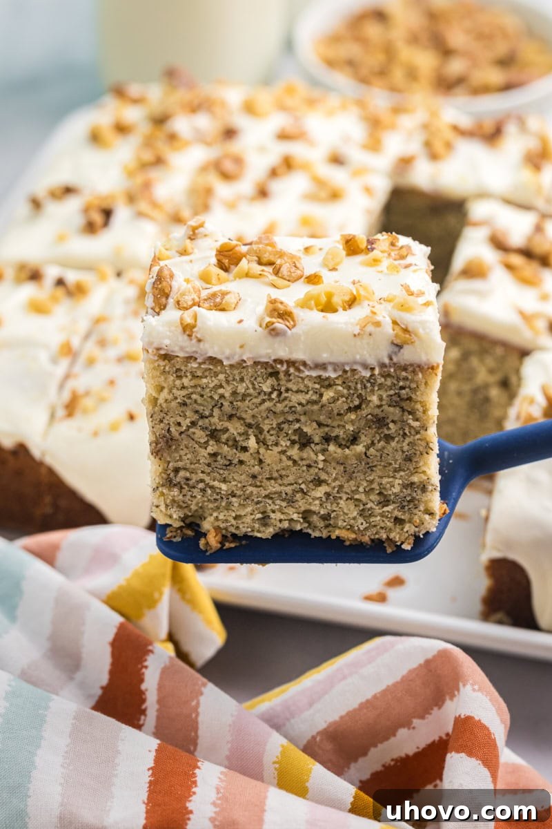 A delectable slice of banana cake with cream cheese frosting, elegantly presented on a cake server, with the remaining frosted cake invitingly blurred in the background.