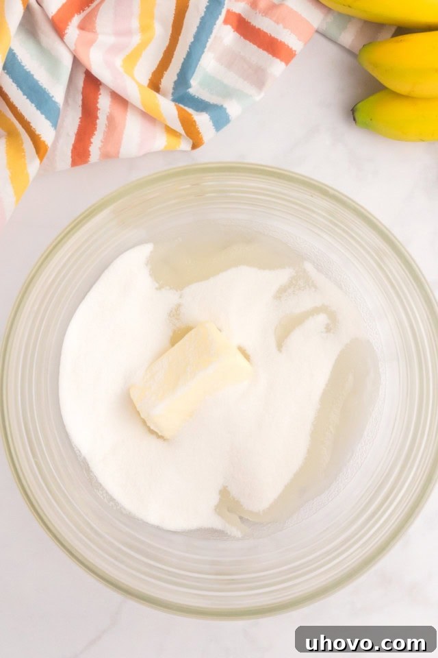 An overhead view of softened butter, canola oil, and granulated sugar in a clear glass mixing bowl, ready to be creamed together.