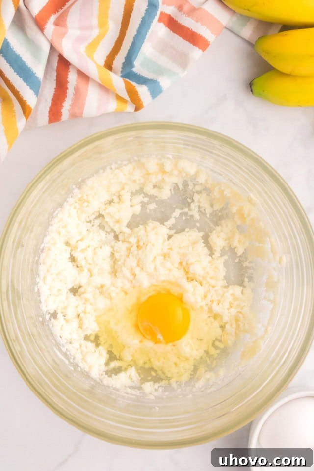 An overhead view of a cracked egg just added to the creamed butter and sugar mixture in a glass bowl, ready for mixing.