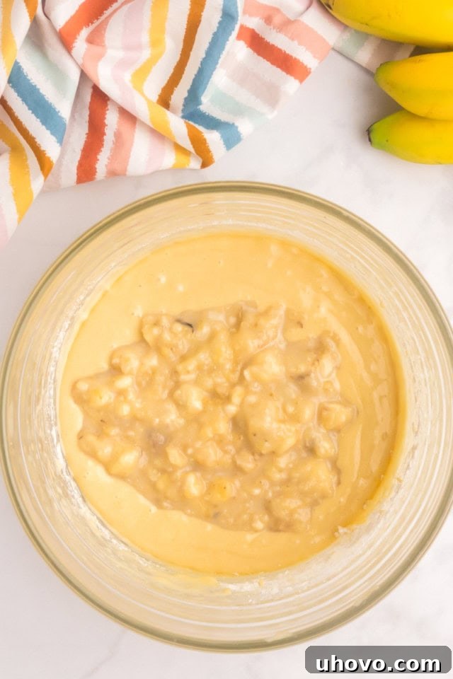 An overhead view of the banana and milk mixture being poured into the partially mixed cake batter in a glass bowl, followed by the remaining flour.