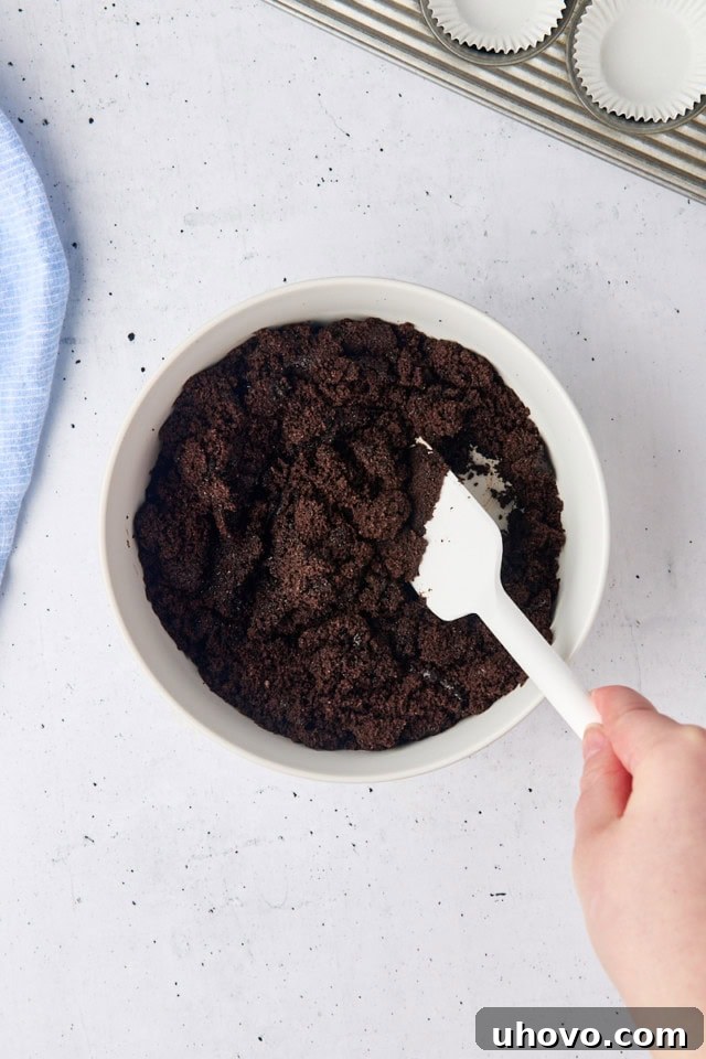 Overhead view of rich Oreo cookie crumbs being mixed with melted butter in a pristine white bowl, forming the base for the cheesecake crust.