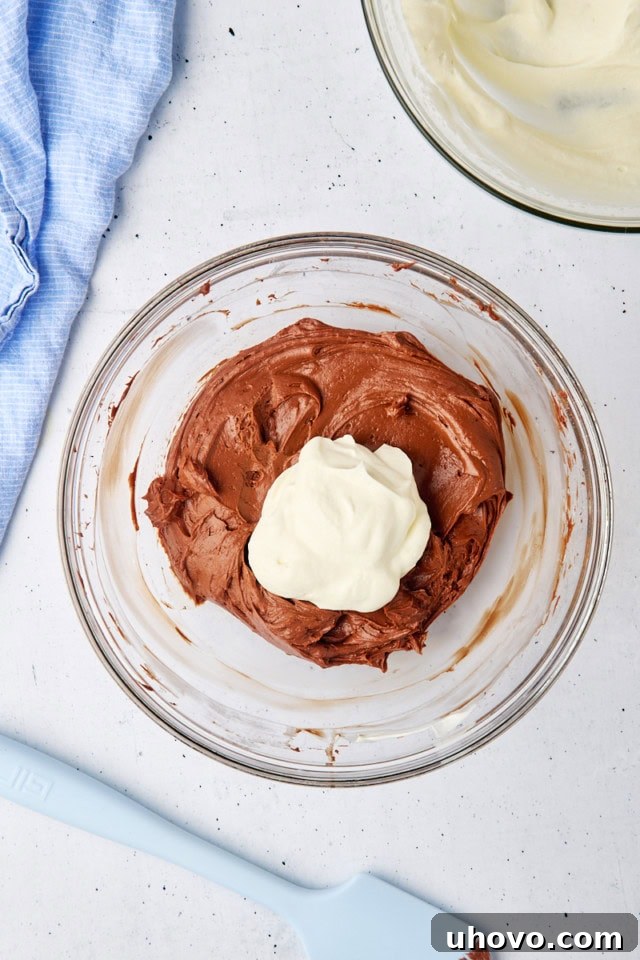Overhead view of a portion of fluffy whipped cream being gently folded into the rich no-bake chocolate cheesecake batter in a glass bowl, ensuring a light texture.