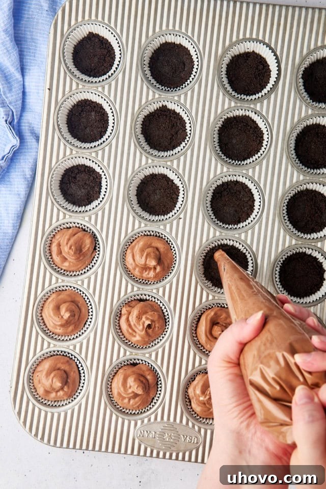 Overhead shot of rich no-bake chocolate cheesecake filling being expertly piped onto the pre-made Oreo cookie crusts in a mini muffin pan, creating perfectly domed tops.
