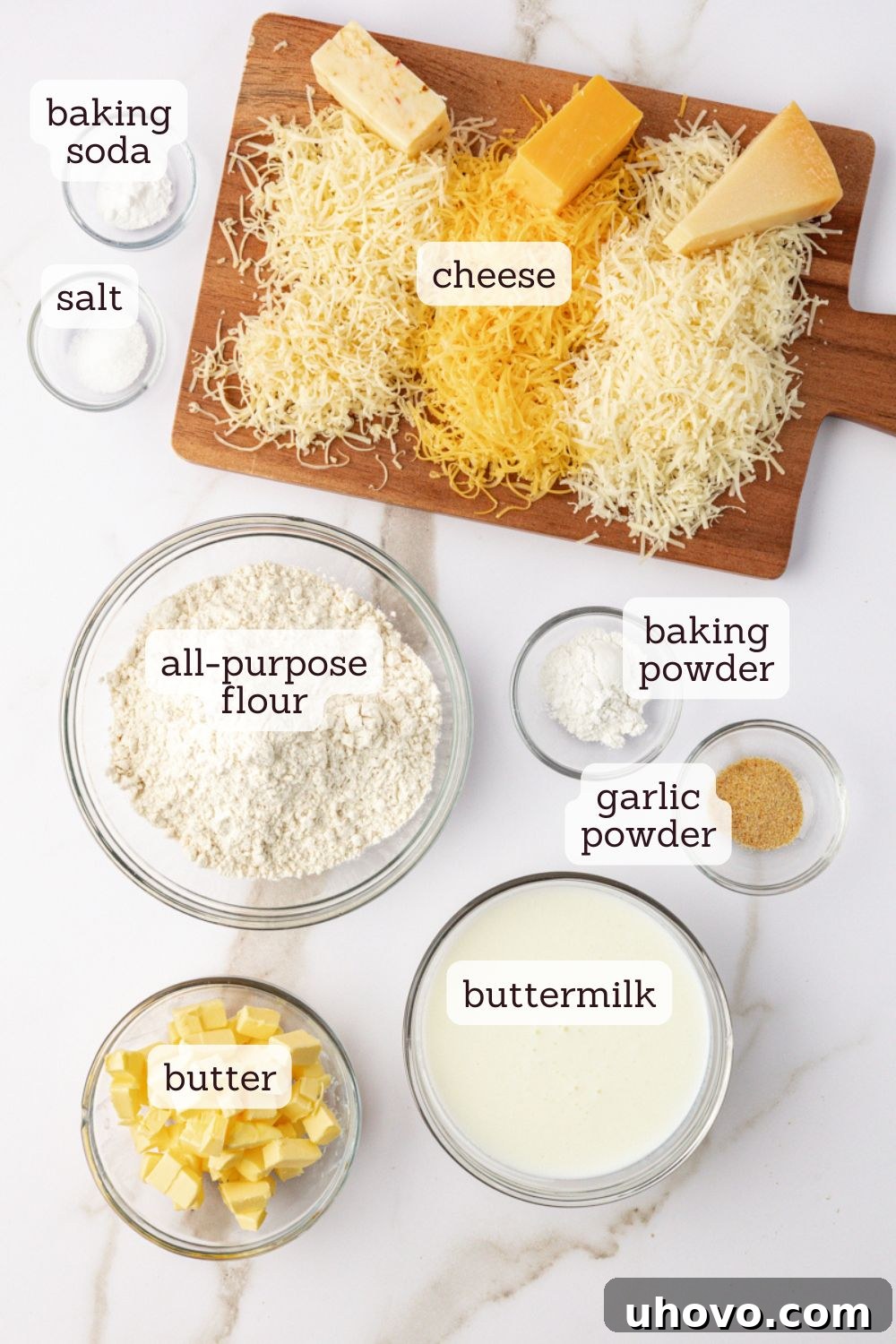 overhead view of key ingredients for three cheese biscuits laid out on a clean surface: flour, butter, buttermilk, and grated cheeses