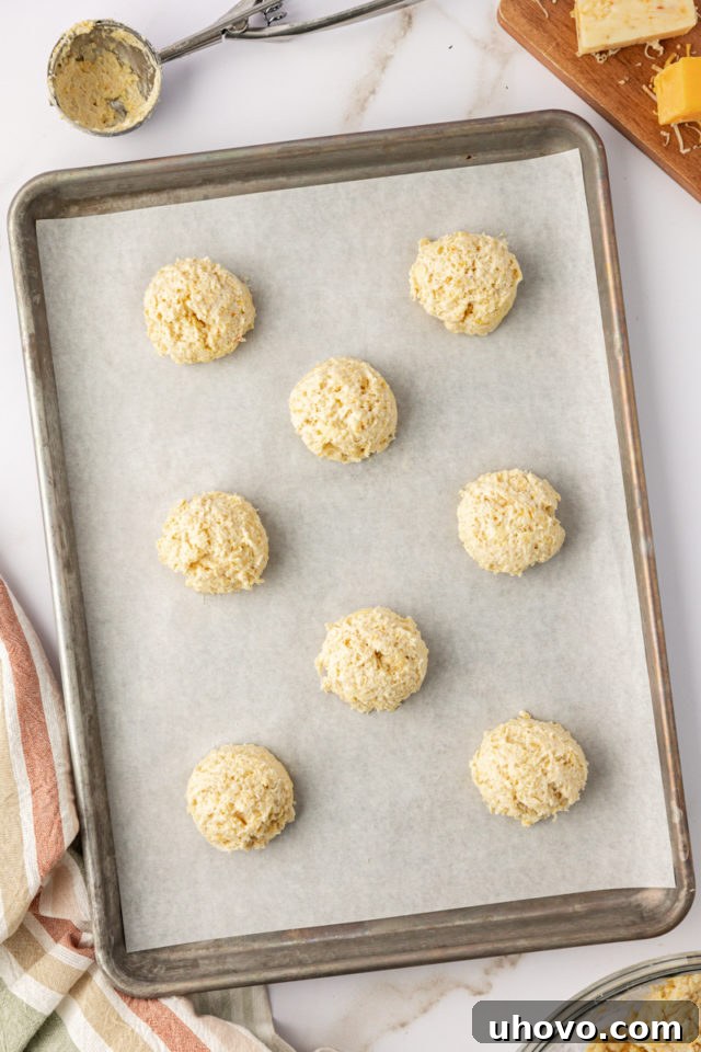 overhead view of three cheese biscuit dough being portioned onto a parchment-lined baking sheet using a cookie scoop, ready to go into the oven